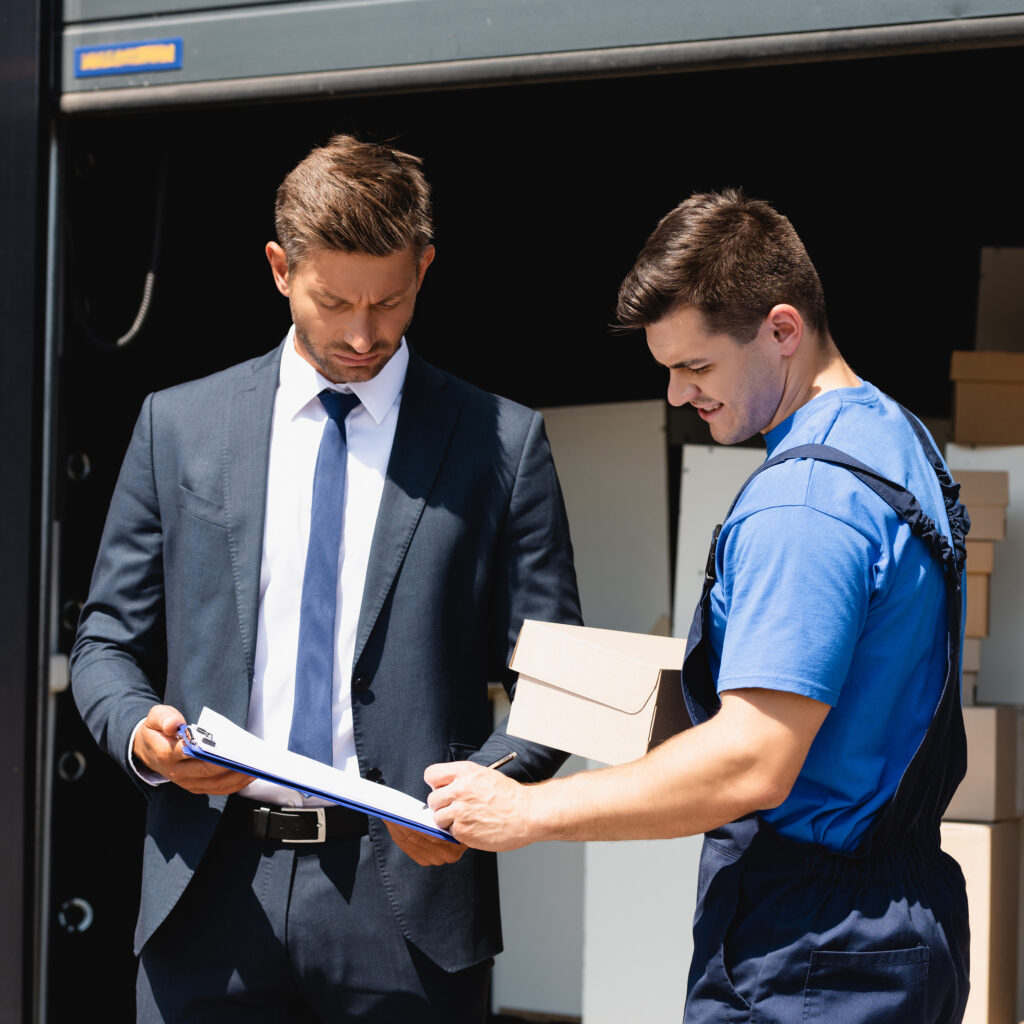 Loader with carton box writing on clipboard near businessman with warehouse at background